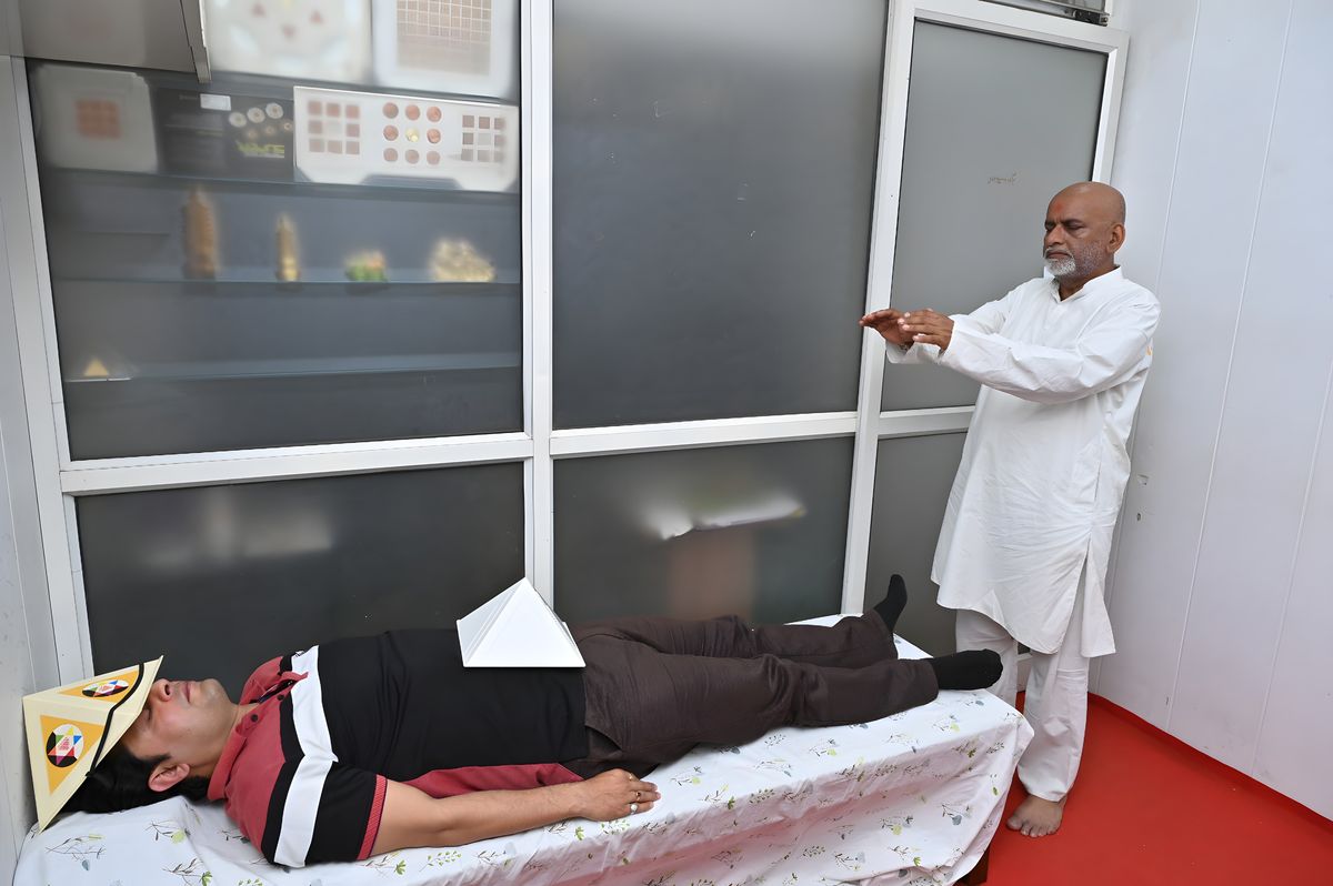 A client lying on a couch with a pyramid cap on his head and a pyramid on his chest while Sanjay Shrivastava heals with open palms